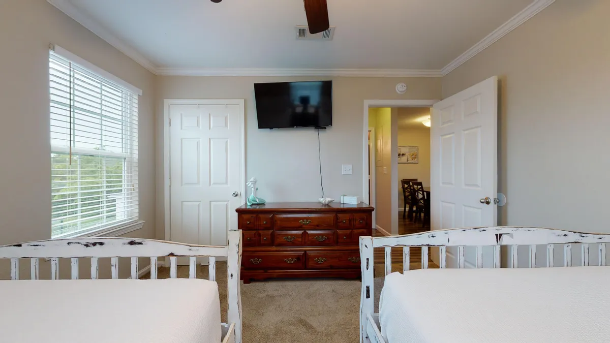 A guest bedroom with two white double beds, a brown dresser, and a television mounted on the wall. The room radiates a comfortable and inviting mood, with ample space for families or friends and clean, coordinated decor.
