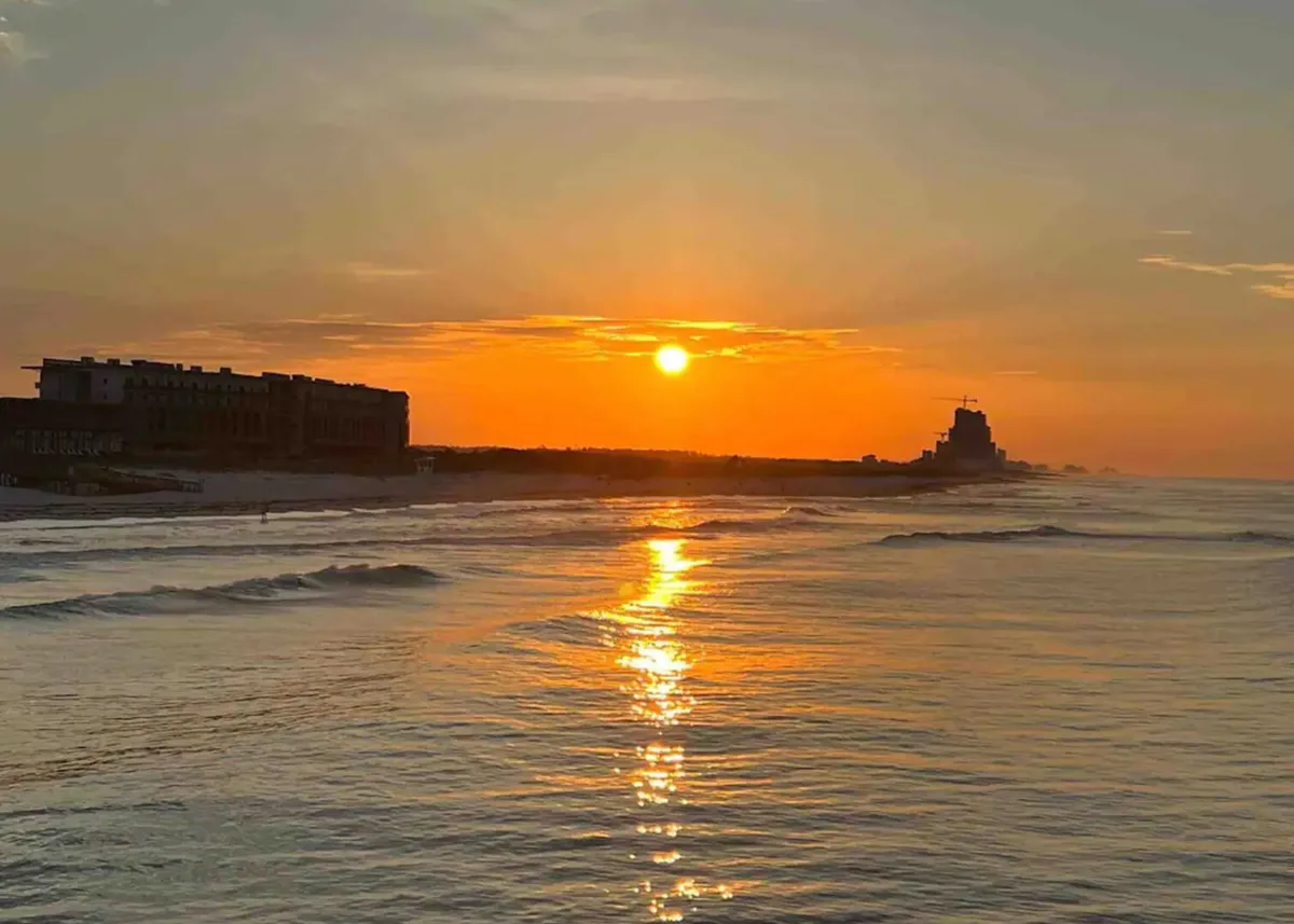 A tranquil beach scene at sunset, with the sun casting golden reflections on the water and distant buildings lining the horizon. The shoreline is calm, inviting visitors to take a peaceful stroll and soak in the beauty of Gulf Shores.