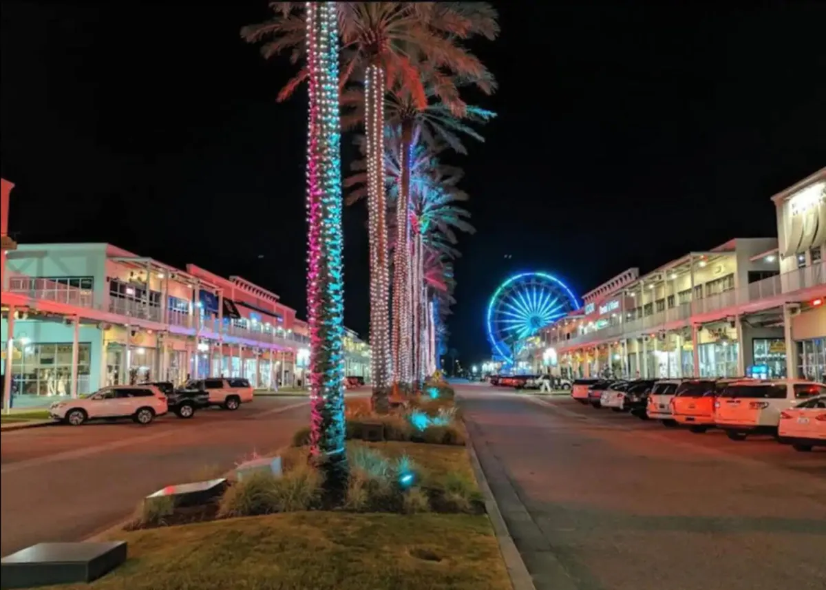 The lively scene includes a Ferris wheel adorned with lights, palm trees, and colorful storefronts, suggesting a festive, amusement-park-like environment perfect for family outings and group fun.