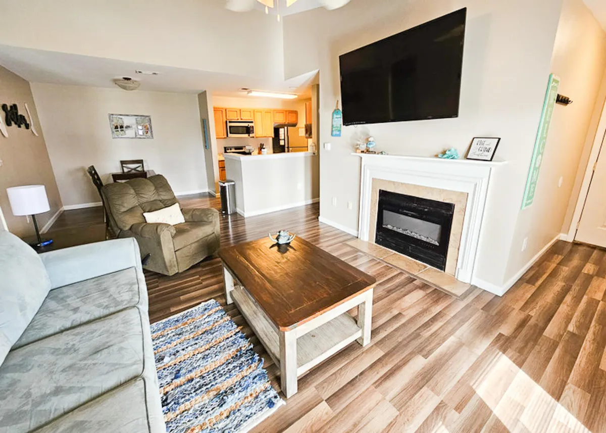 The image features an inviting living room with a plush blue couch, a soft textured rug, a cozy recliner, a stylish wooden coffee table with a decorative centerpiece, and a smart TV mounted above an electric fireplace, creating a warm and comfortable atmosphere.