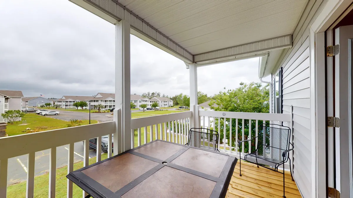 A peaceful private balcony with wooden flooring and white railing, furnished with a metal outdoor table and two chairs. The view overlooks the community’s buildings and landscaped grounds, offering an intimate outdoor extension of the living space.