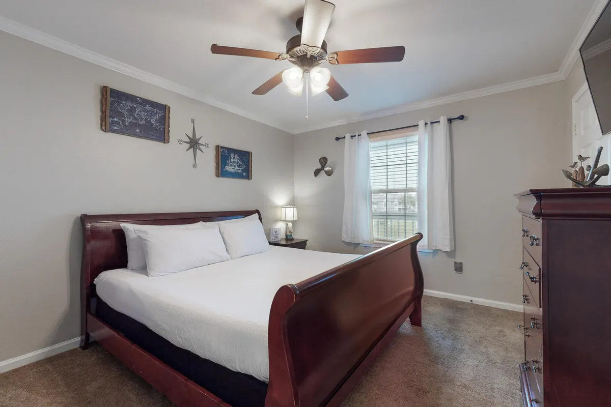 A soothing, well-lit King Suite featuring a wooden sleigh bed with plush white linens, a wooden dresser adorned with decorative accents, a ceiling fan for comfort, and a window with white curtains letting in natural daylight. The overall ambiance is peaceful and inviting, ideal for rest and privacy.