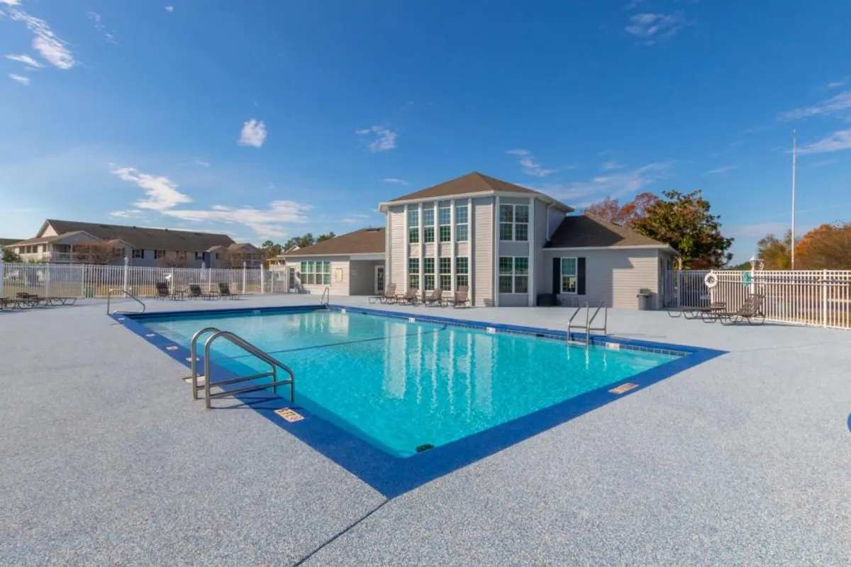 An inviting outdoor swimming pool with crystal-clear water sits beside a modern clubhouse building with large windows. Poolside lounge chairs are arranged around the pool, and a surrounding fence ensures safety and privacy.