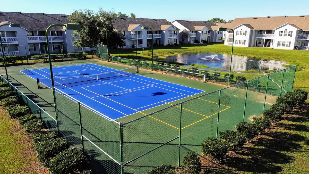 A bright blue tennis court bordered by well-kept green bushes, with a pond and fountain in the background and white residential buildings with balconies nearby. The court is clearly marked, inviting both tennis and pickleball play.
