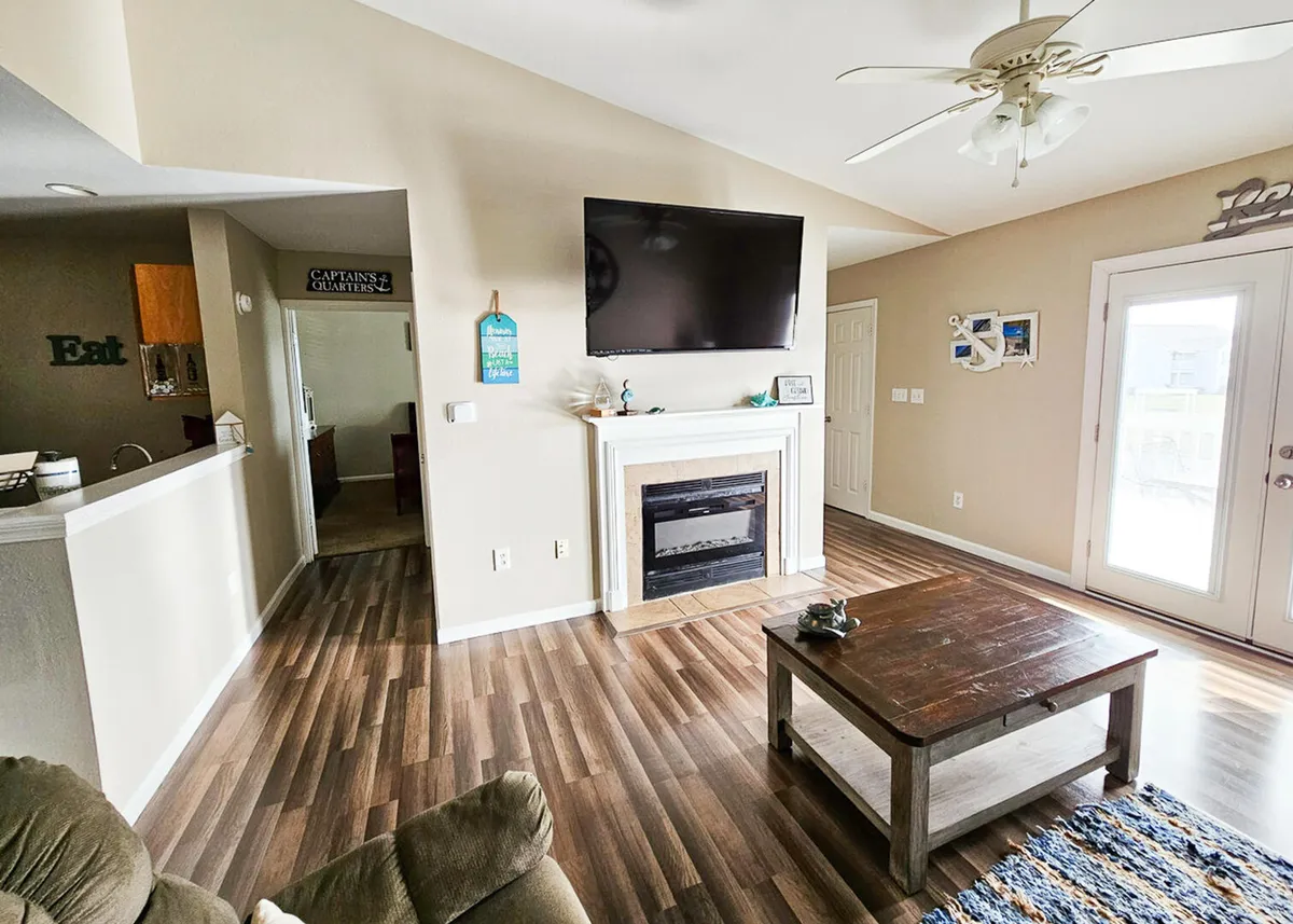 A bright and inviting living room featuring a wall-mounted Smart TV above a decorative fireplace, a stylish wooden coffee table with a decorative centerpiece, and a glass door leading out to a private balcony. The seating arrangement is cozy and ideal for group gatherings or relaxed evenings.