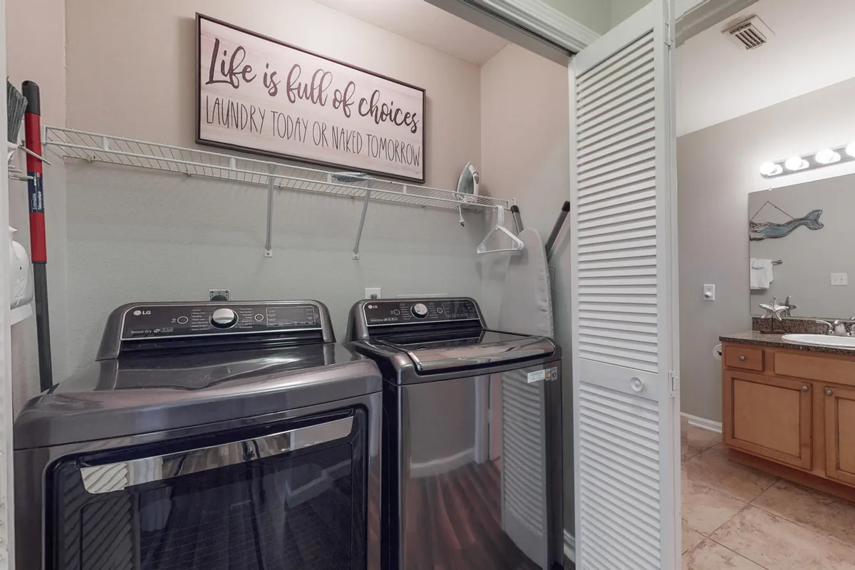 A modern laundry area featuring a washer and dryer set tucked neatly into a closet, with a humorous wall sign about laundry choices adding a touch of personality. The area is bright and practical, with a glimpse of an adjacent bathroom vanity.