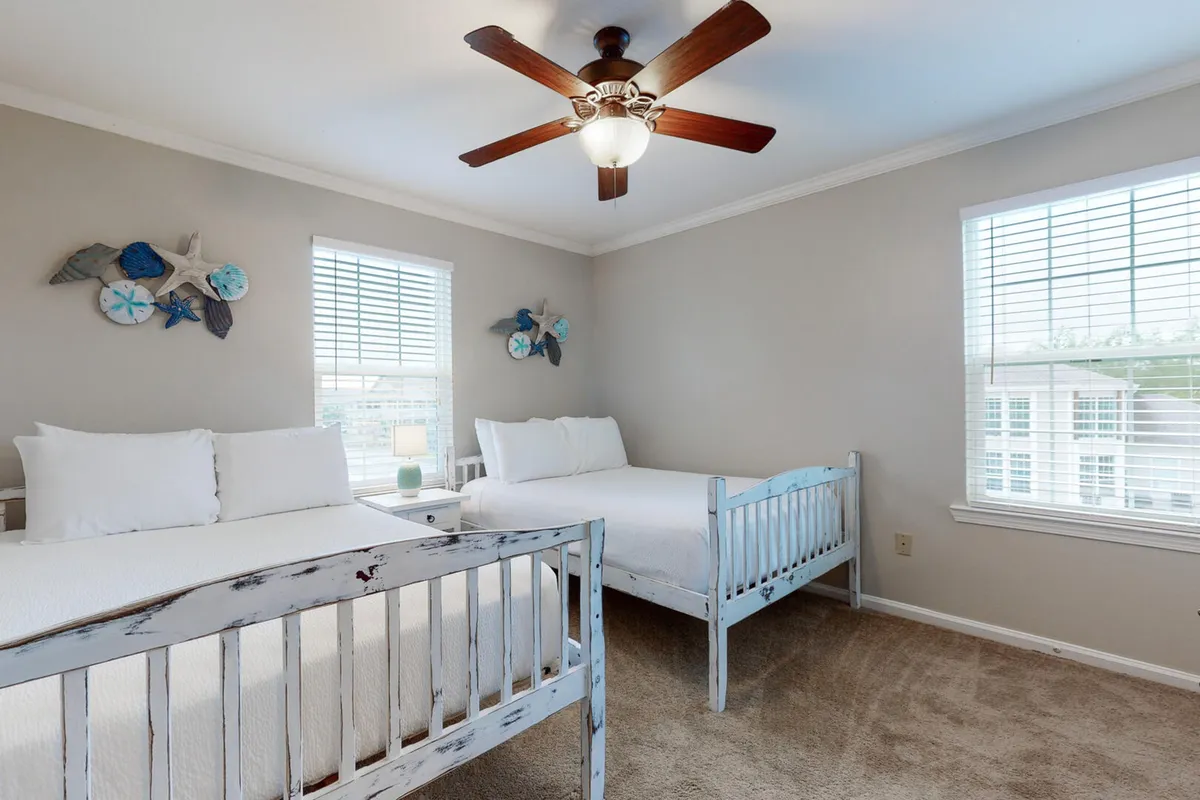 A cozy guest bedroom furnished with two distressed blue wooden beds dressed in crisp white bedding, coastal-themed wall art above the beds, a ceiling fan, and two windows allowing daylight to fill the room. The space is spacious and designed for comfort, perfect for families or friends.