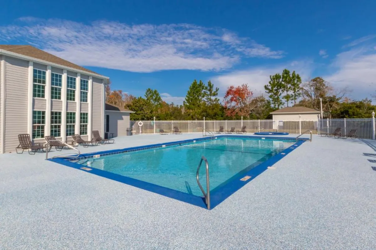 A large rectangular swimming pool with sparkling blue water, surrounded by a spacious deck with multiple lounge chairs for sunbathing. The pool area is enclosed by a white safety fence, and an adjacent building with tall windows completes the inviting atmosphere.