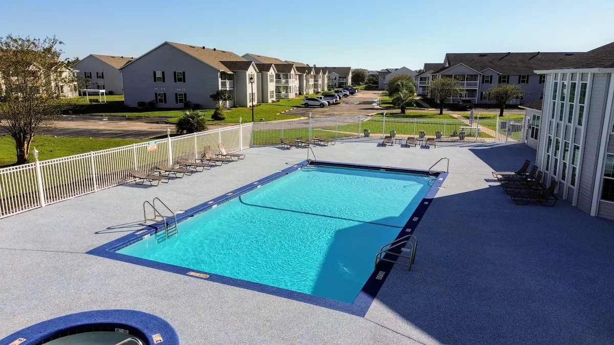 A sunlit outdoor community swimming pool with clear blue water, ringed by neatly arranged lounge chairs and bordered by residential condominium buildings. The pool area is fenced for safety and privacy, creating a welcoming environment for relaxation and socializing.