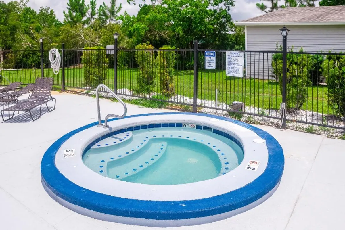 A circular hot tub with a blue and white border, equipped with a metal handrail for safe entry. Lounge chairs are positioned around the area, which is securely enclosed by a black metal fence.