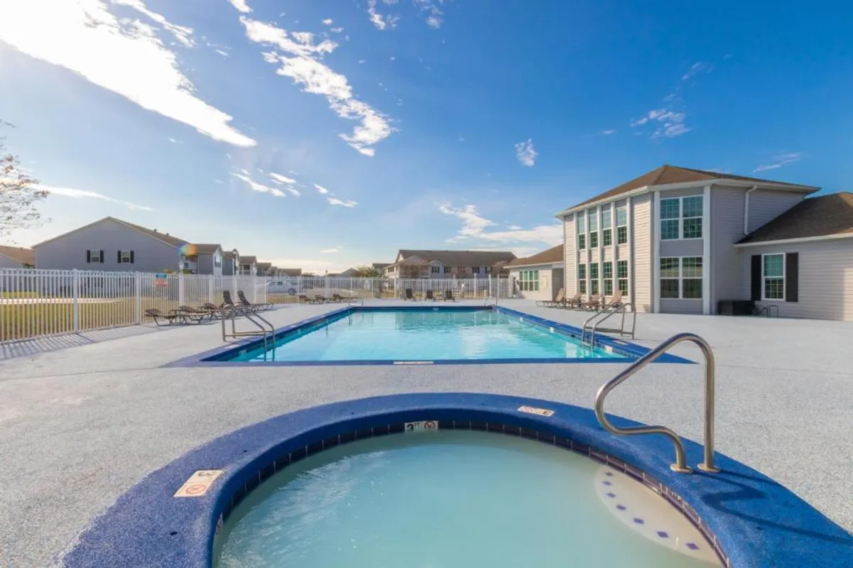 A large outdoor swimming pool and attached hot tub are surrounded by poolside lounge chairs and a community clubhouse building. The area is inviting, perfect for relaxing and unwinding after a busy day at local events or sports competitions.