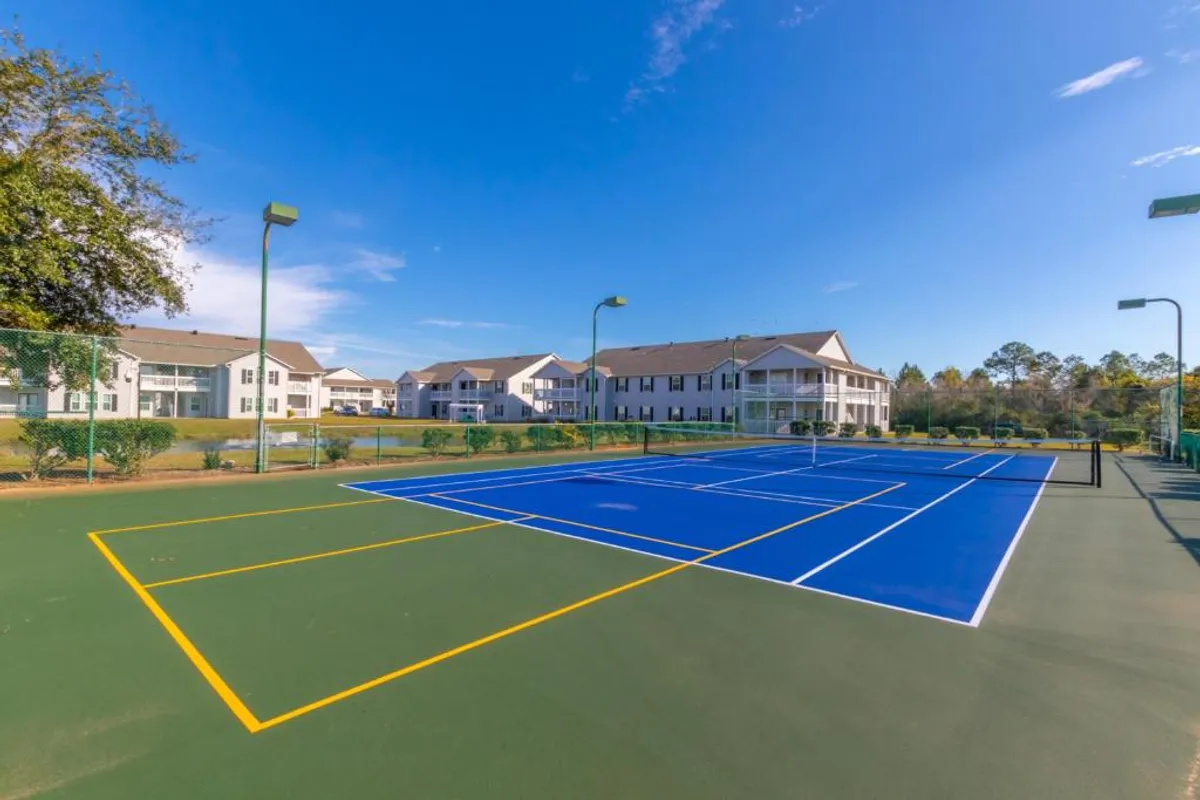 A vibrant blue tennis court is surrounded by greenery and adjacent residential buildings. Tall light poles suggest play can extend into the evening, and the overall environment is active yet inviting, ideal for sports enthusiasts.