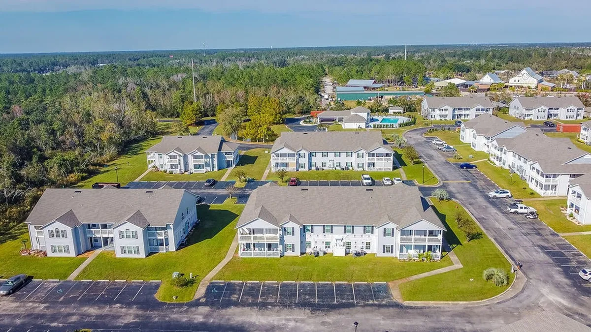 A cluster of white multi-unit residential buildings with private balconies is surrounded by well-maintained lawns, landscaped areas, and marked parking lots. A backdrop of dense forested area gives a sense of privacy and tranquility.