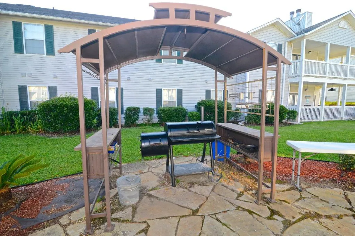 A covered barbecue grill sits on a stone patio surrounded by a green lawn and landscaped shrubbery. Nearby, residential buildings provide a sense of community, making this spot ideal for outdoor grilling and social gatherings.