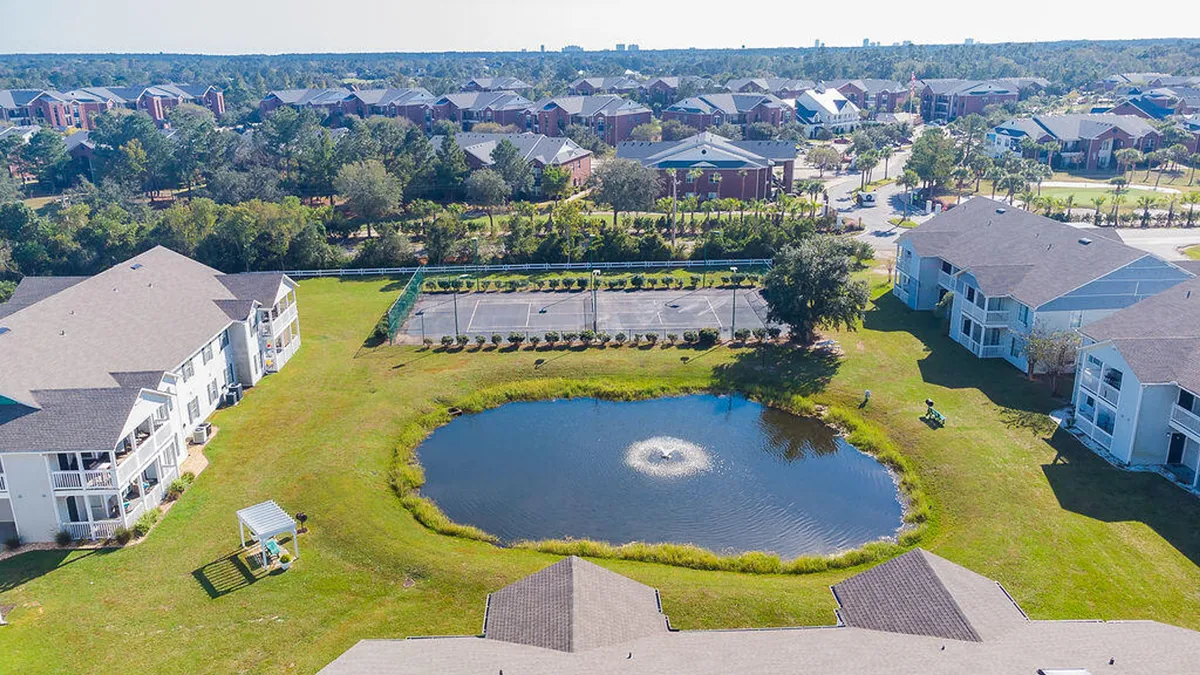 A tranquil pond with a central fountain is framed by well-kept green lawns, tennis courts, and a cluster of condominium buildings. There is also a shaded seating area, creating an inviting, family-friendly atmosphere.