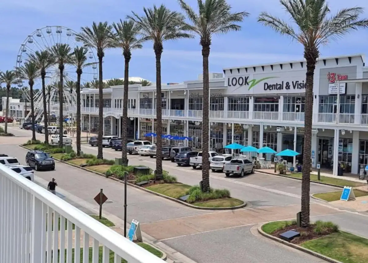 A palm-lined, vibrant street features modern two-story commercial buildings with balconies, blue umbrellas shading outdoor seating, and a large Ferris wheel in the distance. The scene is lively, inviting, and perfect for family exploration.