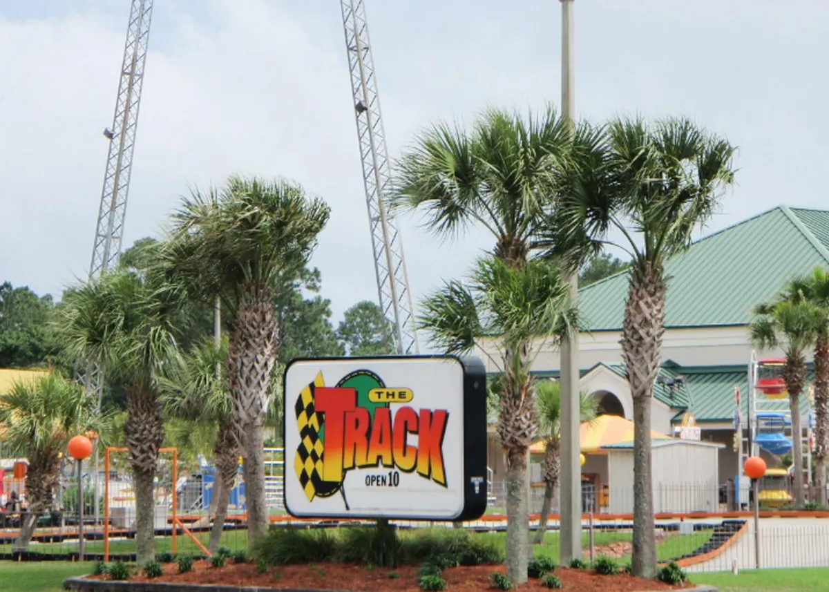 The entrance to The Track amusement park is showcased, with a prominent racing flag sign, palm trees lining the area, and colorful amusement rides visible in the background. The landscaping is neat, and the overall scene is bright and full of energy, suggesting a vibrant family-friendly attraction.