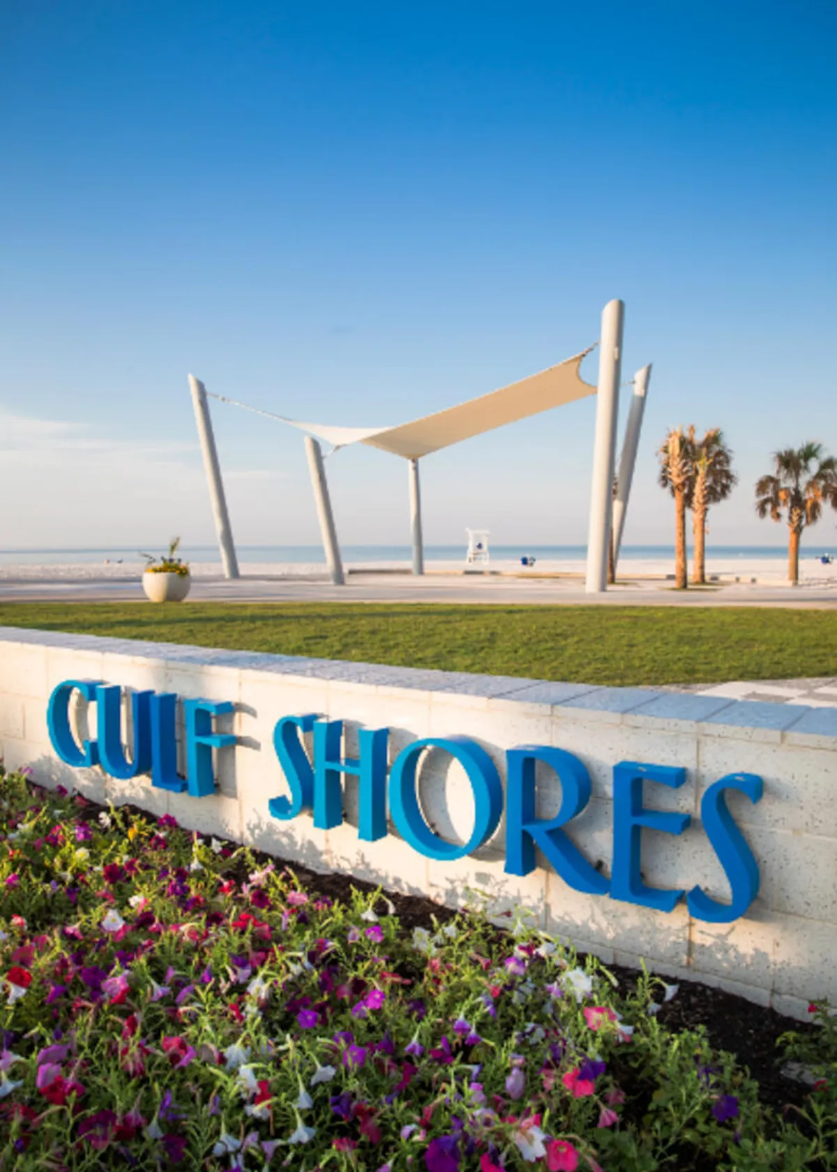 A stylish blue 'Gulf Shores' sign anchors a landscaped entrance with palm trees and a modern white shade structure. Vibrant flowers bloom in the foreground, and the beach is just beyond, inviting visitors to enjoy the sun and sand.